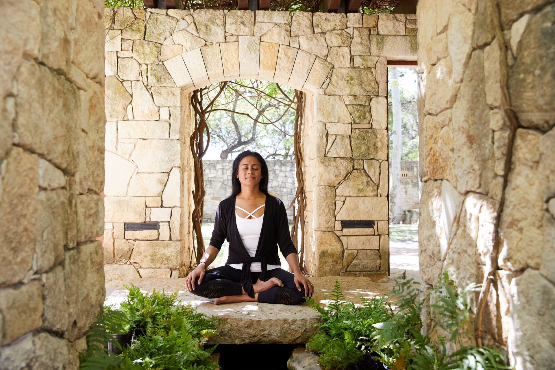 Woman sitting outside in garden in front of stone wall doing yoga